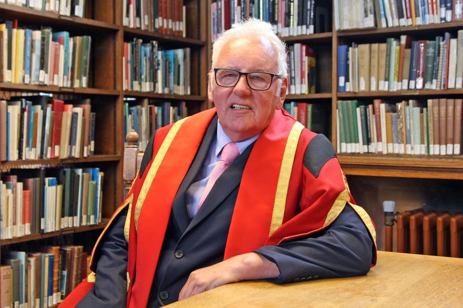 Image of a man wearing glasses and red and gold graduation robe. His right arm leans on the table before him. There are bookshelves in the background.