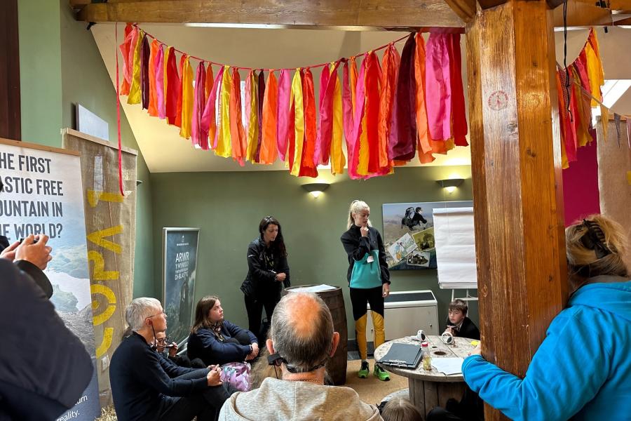Discussion taking place in an informal setting with Casi Wyn (Poet) talking to a room of attendees, some of which are standing and some sitting down. Colourful banners hang from the ceiling to celebrate the launch of the event